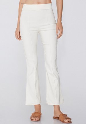 Pantaloni bianchi a zampa d'elefante con una texture di tessuto liscia, vita alta e sottili cuciture verticali; abbinati a sandali scivolati marroni con texture.