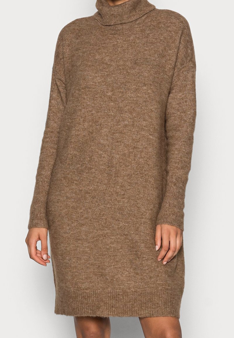 Femme portant une longue robe-pull marron, duveteuse avec un col roulé, des manches longues et des poignets et un bas côte.