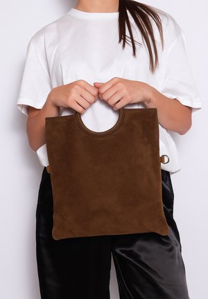 Bruine suède tote bag met een afgeronde handgreep, gekenmerkt door een gladde textuur en een minimalistisch ontwerp, vastgehouden door een persoon die een wit shirt draagt.