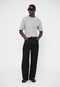 Sivá tričko s krátkym rukávom, čierne široké nohavice a čierne topánky. Modelka má na sebe veľké čierne slnečné okuliare; neutrálne pozadie.