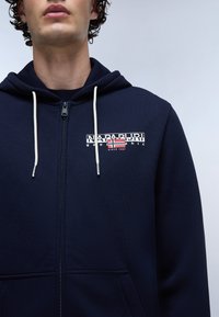 Felpa con zip di colore blu navy, dotata di stringhe, presenta una stampa del logo con una bandiera, realizzata in tessuto morbido, include tasche frontali e una finitura testurizzata.