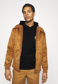 Brown suede jacket with a furry collar, zip closure, side pockets, and knit cuffs. Worn over a black hoodie, with a simple design.