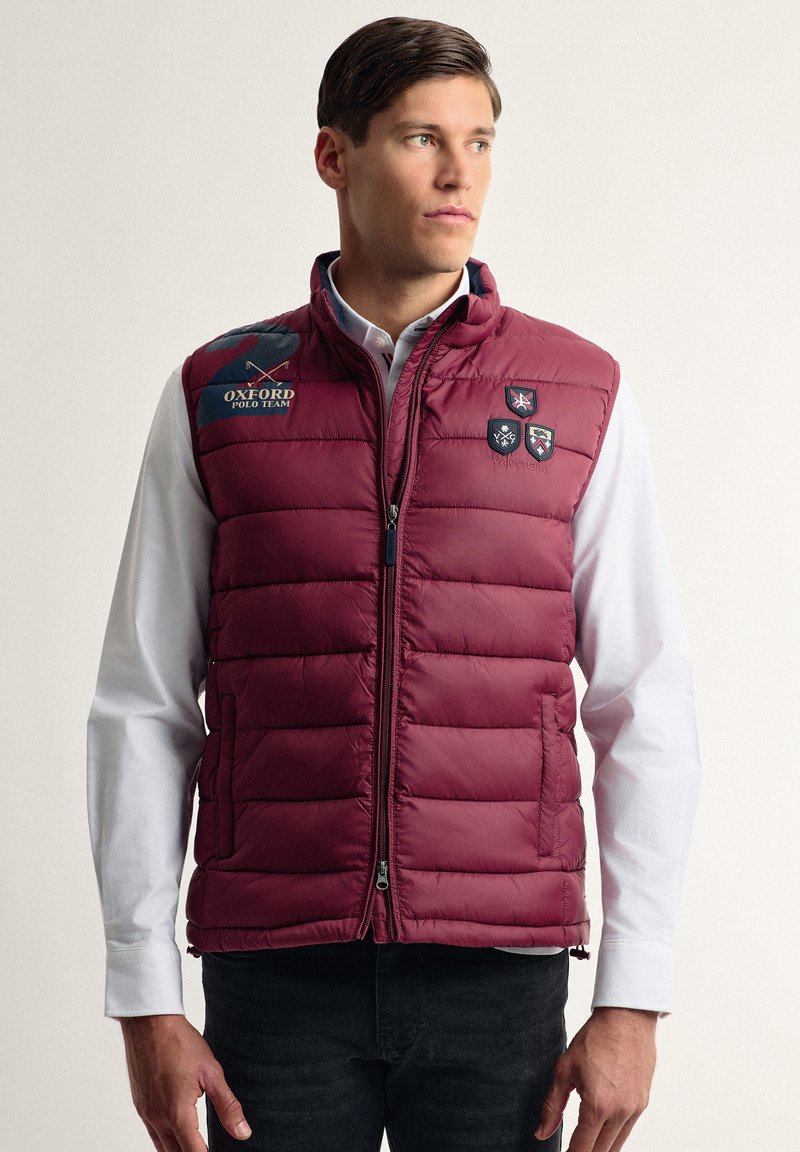 Chaleco acolchado burdeos con cremallera frontal, que presenta costuras horizontales, parches con emblemas y bolsillos laterales. Llevado sobre una camisa blanca.