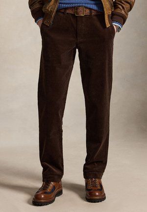 Homme portant un pantalon en velours côtelé marron foncé, une ceinture en cuir marron, un pull bleu, une veste marron et des bottines à lacets marron sur fond neutre.