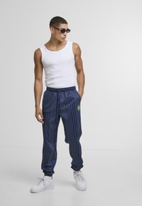 Pantaloni navy a righe con polsini elasticizzati e cordino, abbinati a una canotta bianca e a sneaker bianche. Gli accessori includono occhiali da sole scuri.