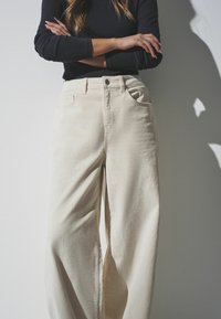 Beige wijde broek gemaakt van gestructureerd katoen, met een sluiting aan de voorkant met knoop, riemlussen en zijzakken.