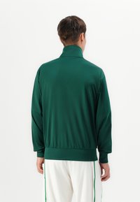 Grønn sportsjakke med høy krage, laget av glatt stoff; har en tettsittende midje og lange ermer. Enkel design, ingen synlige mønstre.