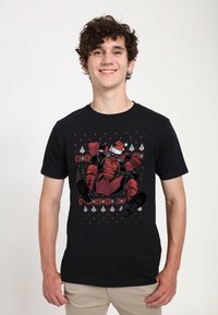 Svart bomull t-shirt med en röd superhjälte i tomteluva, omgiven av festliga mönster, inklusive träd och snöflingor.