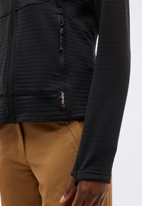 Približnji posnetek osebe, ki nosi črno Polartec jakno z zadrgo in rjave hlače, prikazuje stranski žep in detajle na rokavu jakne.