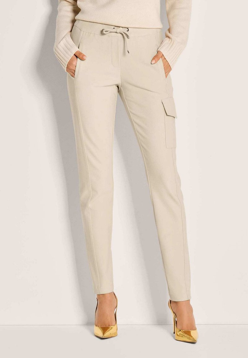 Beige slim-fit broek met zijzakken en een cargozak, gecombineerd met gouden high heels en een crèmekleurige lange mouw top.