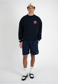 Homme debout portant un sweat-shirt bleu marine avec un logo rouge, un short assorti, une casquette blanche, des chaussettes blanches avec écriture et des mocassins noir et blanc sur fond blanc.