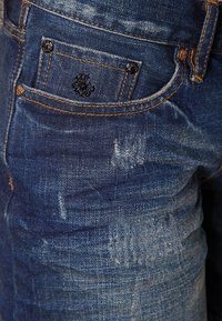 Jeans de mezclilla azul oscuro con una textura desgastada, que presentan un pequeño bolsillo trasero y costuras en contraste. Incluyen un logo bordado en negro.