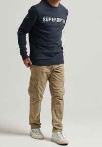 Navy crewneck sweatshirt with white "SUPERDRY" text, paired with beige cargo pants featuring multiple pockets and white sneakers.