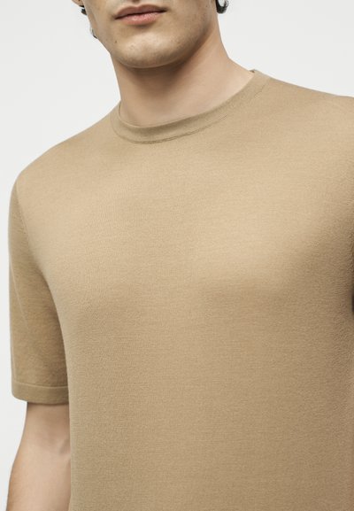 Κοντινό πλάνο ενός ατόμου που φορά στενά μπεζ πλεκτό μπλουζάκι με κοντά μανίκια και στρογγυλή λαιμόκοψη, με λευκό απλό φόντο.