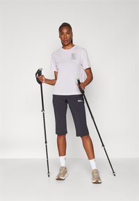 Tricou de culoare lavandă deschisă cu buzunar pe piept și pantaloni scurți gri de drumeție. Modelul ține bețe de trekking și poartă adidași bej și șosete albe.