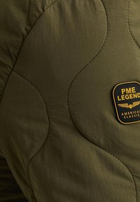 Olivgrüner gesteppter Stoff mit geschwungenen Nähmustern. Verfügt über ein rechteckiges schwarzes und gelbes Logo-Label mit der Aufschrift "PME LEGEND."