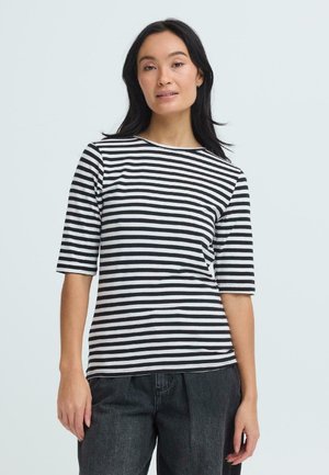 Kobieta o długich ciemnych włosach, ubrana w czarno-biały koszulę w poziome paski oraz ciemne spodnie, stojąca na tle jednolitego jasnego tła.