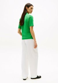 Zielona koszula z krótkim rękawem, wykonana z gładkiego materiału, w połączeniu z luźnymi białymi spodniami i czarnymi mokasynami. Model stoi tyłem do aparatu.