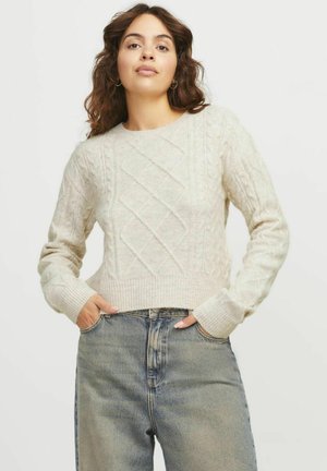 Maglione all'uncinetto color beige chiaro con motivo a treccia, scollo rotondo e polsini a coste. Abbinato a jeans a vita alta blu sbiaditi.