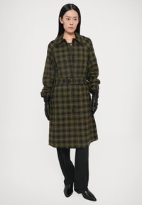 Mustvalge kollase ruudulise mustriga kleit vöökohal vööga, ees nööbitav ja pika varrukaga, sobitatud mustade kinnaste ja kohandatud mustade pükstega.