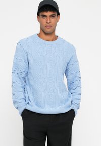 Ανοιχτό μπλε πλεκτό πουλόβερ με υφι323σμένες καλωδιωτές υφές, στρογγυλή λαιμόκοψη και ρίγες στα μανίκια και το τελείωμα. Συνδυασμένο με μαύρο παντελόνι.