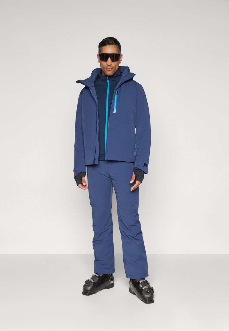 Veste de ski marine avec capuche et zip turquoise, associée à un pantalon marine et des bottes de ski noires. Comprend des manches longues avec des trous pour les pouces.
