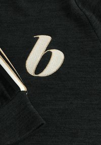 Zwarte stof met een zachte textuur, voorzien van een grote geborduurde "b" in crème-kleur en witte en zwarte strepen aan de manchetten.