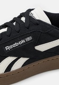 Svart mockasinsneaker med vita detaljer, försedd med en texturerad brun gummisula, vadderad krage och "Reebok"-logotyp på sidan.