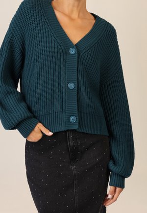 Personne portant un cardigan en grosse maille bleu sarcelle boutonné et une jupe noire avec de petits clous argentés, fond beige.