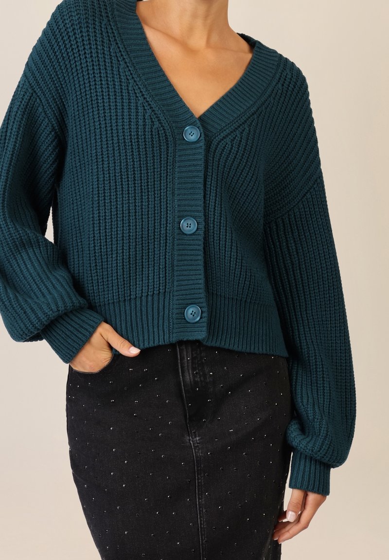 Personne portant un cardigan en grosse maille bleu sarcelle boutonné et une jupe noire avec de petits clous argentés, fond beige.