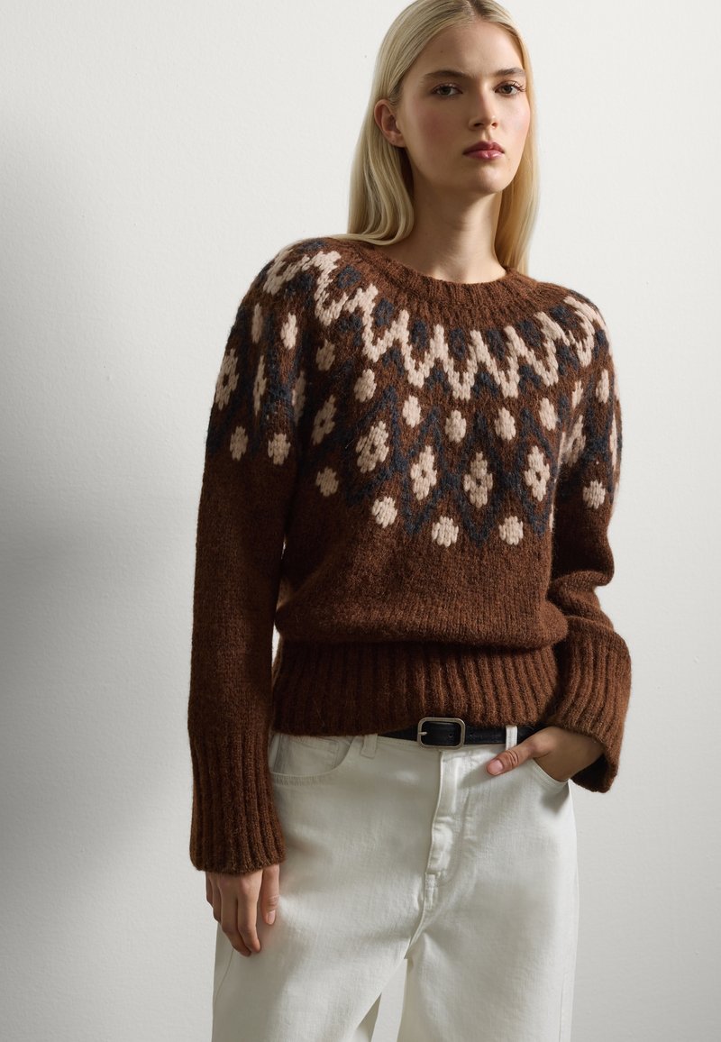 Suéter marrón de punto con un yugo de diseño en beige y azul, con puños y dobladillo acanalados. Combinado con unos jeans blancos de cintura alta.