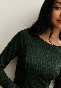 Donna che indossa un abito lungo a maniche verdi scuro con stampa leopardata nera, in piedi contro un muro beige chiaro.