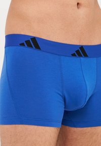 Mėlynos boxer kelnaitės pagamintos iš švelnios, tampos medžiagos. Yra platus mėlynas liemuo su juodais Adidas logotipo akcentais. Matomas siūlių apdorojimas.