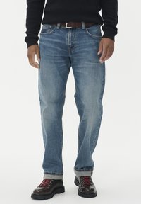 Vyras, apsirengęs mėlynomis džinsinėmis kelnėmis, rudos odinės diržo, juodu megztiniu ir tamsiai rudais batais su raudonais raišteliais, ant švaraus balto fono.