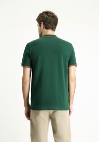 Polo verde con colletto piatto, maniche corte e dettagli a strisce arancioni sui bordi delle maniche. Tessuto morbido, design aderente.