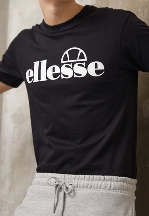 Osoba ubrana w czarną koszulkę z białym logo "ellesse" i grafiką w kształcie półkola, w połączeniu z jasnoszarymi spodnimi dresowymi z sznurkiem.