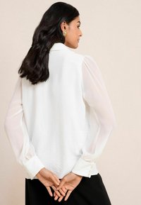 Blusa bianca con corpo testurizzato e maniche a pallone trasparenti. Presenta polsini con bottoni e un colletto semplice. Indossata con le mani incrociate dietro la schiena.