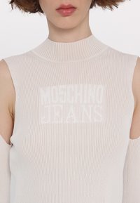 Ribota, šviesiai smėlinė be rankovių palaidinė su aukštu kaklu. Yra pjūviai pečių srityje ir balta "Moschino Jeans" logo krūtinėje.