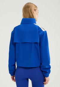 Blauwe sportjacket met hoge kraag, verlengde achterzoom en witte accentstrepen op de schouders, voorzien van elastische manchetten voor een strakke look.