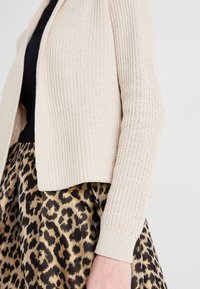 Beige strikket cardigan med ribbestruktur, kombinert med et svart og gull leopardmønstret skjørt med en glansfull overflate.