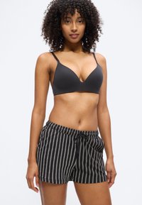 Γυναίκα με σγουρά μαλλιά, ντυμένη με μαύρο σουτιέν και σορτς με μαύρες ρίγες, στέκεται μπροστά σε ένα απλό ανοιχτό γκρι φόντο.