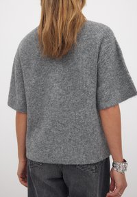 Pilkas megztinis su laisvu siluetu, trumpomis rankovėmis ir švelnia tekstūra. Turi nusmukusį ilgio ir išraižytą raštą. Derinamas su tamsiai pilkais džinsais.