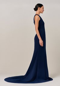 Temno modra obleka s prilegajočo silhueto, okroglim izrezom in vidnimi stranskimi gumbi. Ima dolgo vlečko za dodatno eleganco.