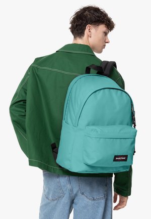 Turkos ryggsäck med rundad design, framficka och svart logotyp. Materialet verkar slätt, kombinerat med en grön jacka och jeans.