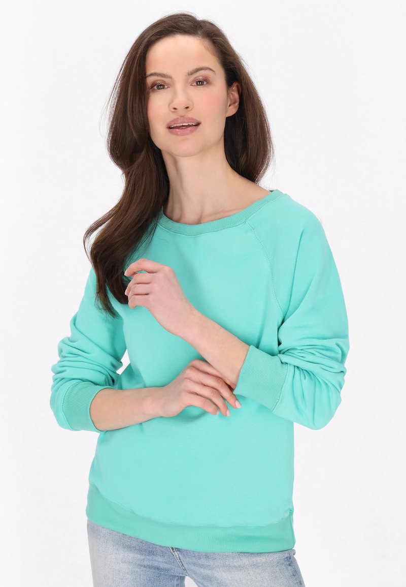 Felpa verde menta con ampio collo a giro e maniche lunghe a raglan, con polsini a coste, indossata sopra jeans azzurri. Texture liscia.