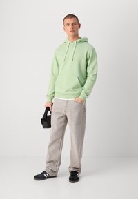 Haut à capuche vert clair avec une poche avant, associé à un jean gris clair. Le mannequin tient un petit sac noir et porte des baskets noir et blanc.