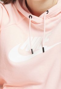 Rosa Nike-hoodie med en vit logotyp, med dragsnören med svarta ändar, mjukt tyg och avspänd passform.