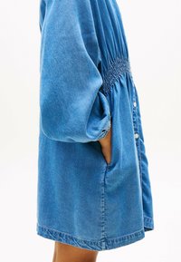 Vestido de mezclilla con cintura fruncida, mangas largas sueltas, bolsillos y cierre de botones en el frente. El color es un azul medio con un ligero brillo.