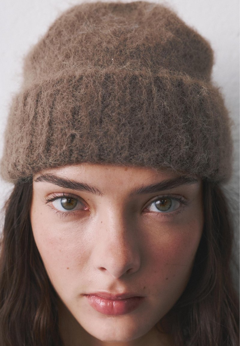 Jeune femme aux yeux noisette portant un bonnet en laine brun duveteux, regardant directement la caméra avec une expression neutre.