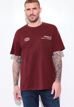 Homme aux cheveux courts et tatouages sur les deux avant-bras, portant un t-shirt bordeaux avec un texte blanc et un jean bleu, debout devant un fond blanc.